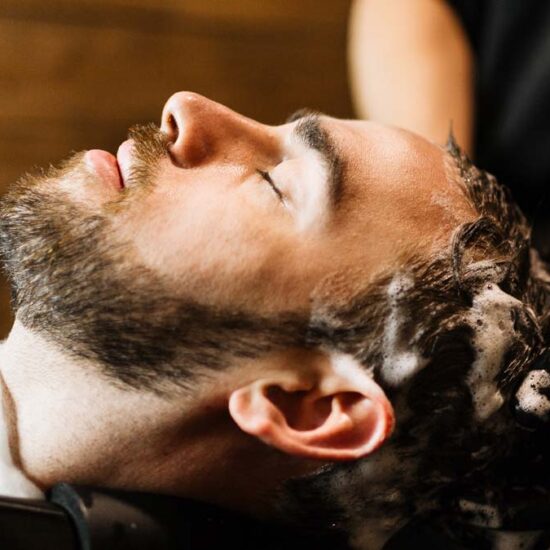 A man with a beard leans back in a salon sink to have his hair shampooed by hands wearing black gloves.