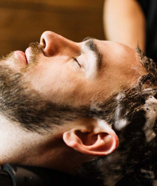 A man with a beard leans back in a salon sink to have his hair shampooed by hands wearing black gloves.