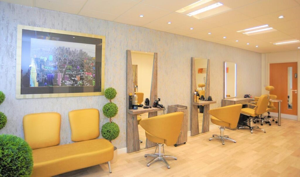 Our modern salon waiting area and styling stations featuring mustard yellow seating, large wooden-framed mirrors and a framed cityscape photograph on the wall.