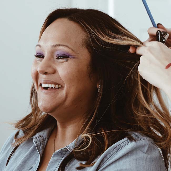 A smiling middle aged woman with brown hair laughs while having her hair styled. She is wearing bright purple eyeshadow.