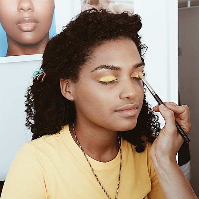 A makeup artist applies bright yellow eyeshadow to a young woman wearing a yellow t-shirt and gold necklace.