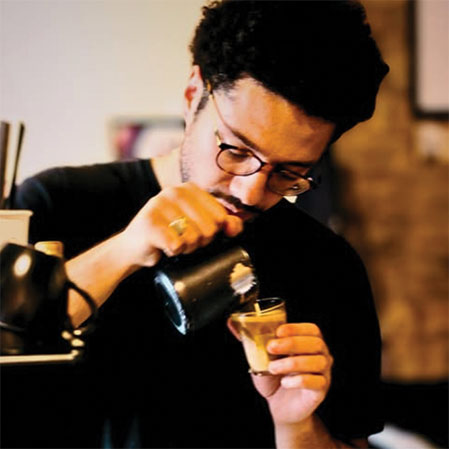 A man wearing glasses and a black shirt carefully pours frothed milk from a black pitcher into a small glass of espresso to create latte art.