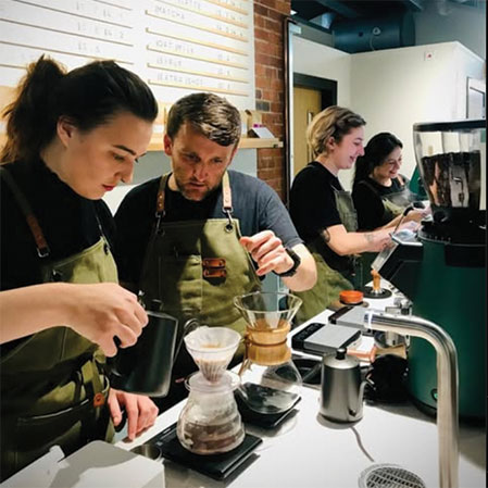 Four Crosby Coffee trainee baristas, all wearing aprons, work behind a busy coffee shop counter, preparing coffee and using a tablet for orders.