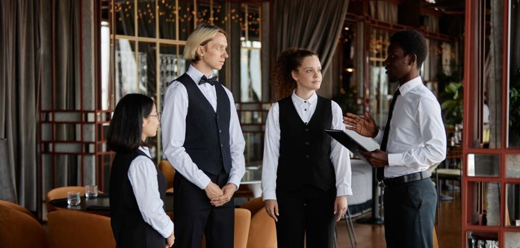 Four restaurant staff members in uniform are gathered indoors; one is holding a clipboard and briefing the team.