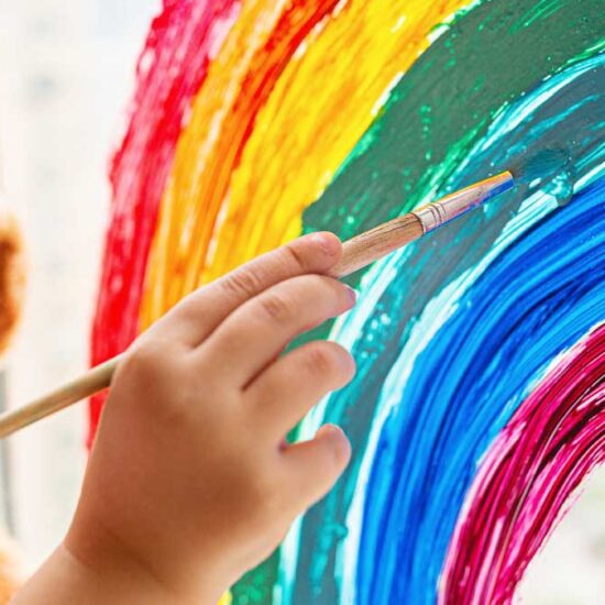 A child's hand using a paintbrush to paint a bright rainbow directly onto a glass window, with a blurred brown teddy bear sitting nearby.