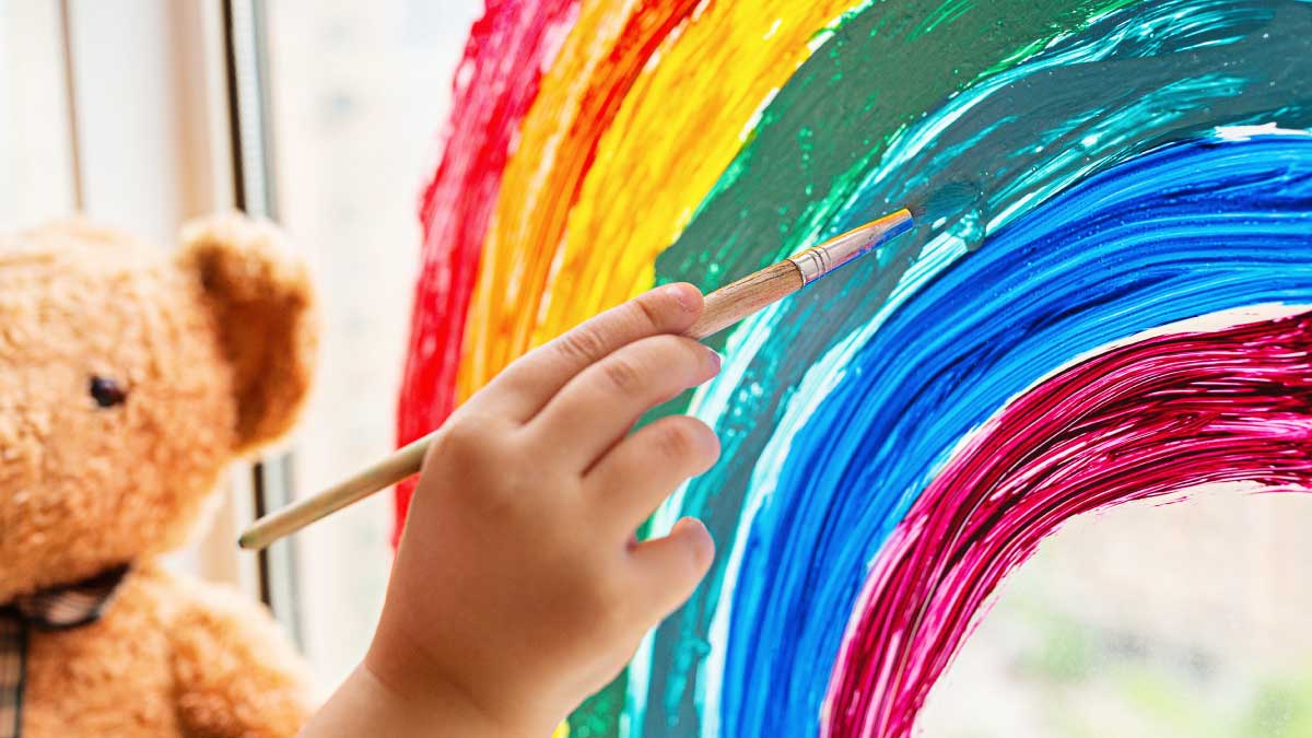 A child's hand using a paintbrush to paint a bright rainbow directly onto a glass window, with a blurred brown teddy bear sitting nearby.