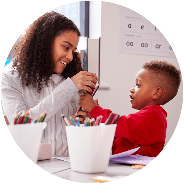 A smiling woman hands coloured pencils to a young child who is excitedly reaching for them during an activity at a table.
