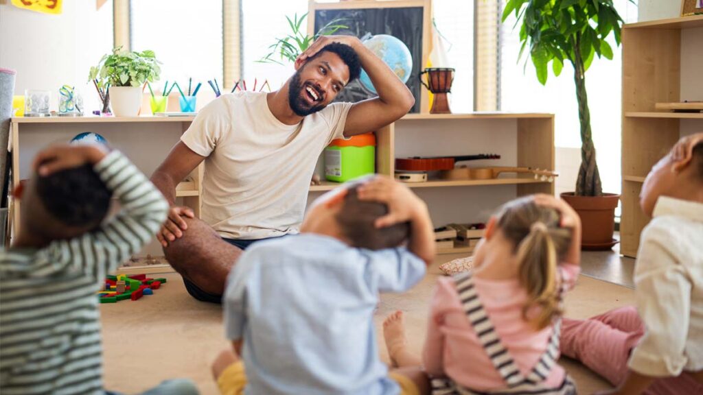 A male classroom assistant leading a group of young children in a stretching activity on a classroom floor.