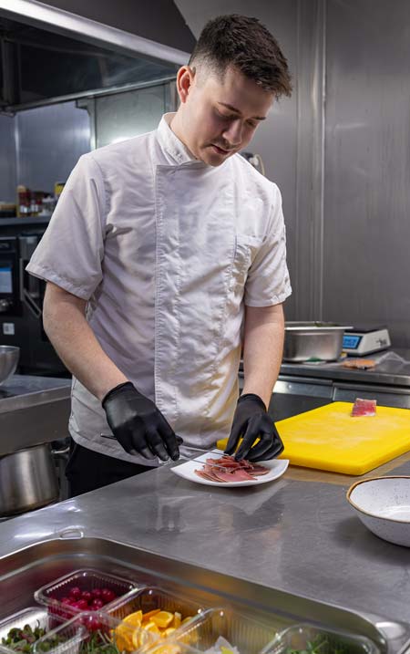 A male chef wearing a white jacket and black disposable gloves arranging slices of sliced meat onto a small white plate in a stainless steel commercial kitchen.