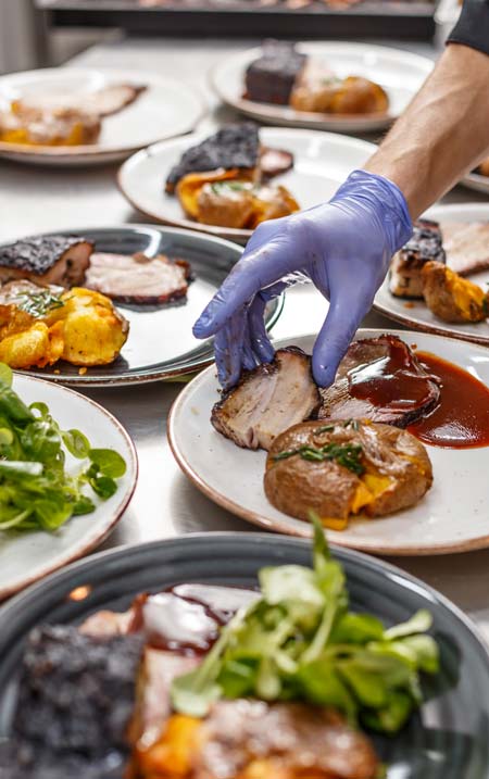 A hand wearing a blue disposable glove carefully placing a slice of barbecued meat onto a plate next to a baked potato and sauce.