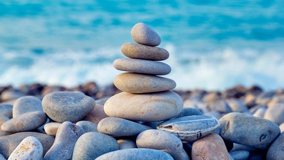 A perfectly balanced cairn of smooth stones on a pebble beach with blue water and waves in the background.