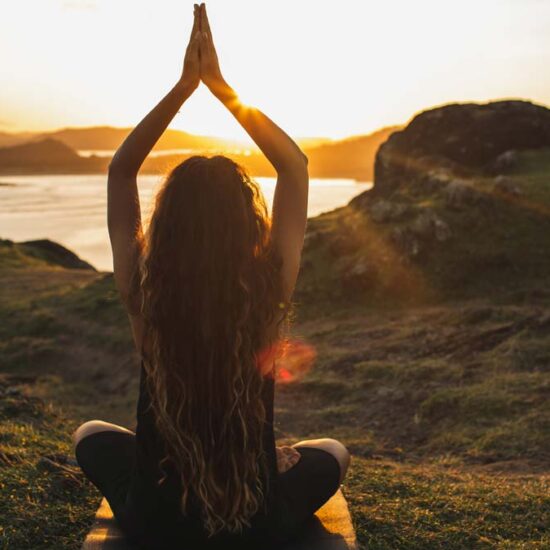 A woman with long wavy hair is sitting in a cross-legged yoga pose on a grassy hill overlooking water, hands raised above her head against a bright sunrise.