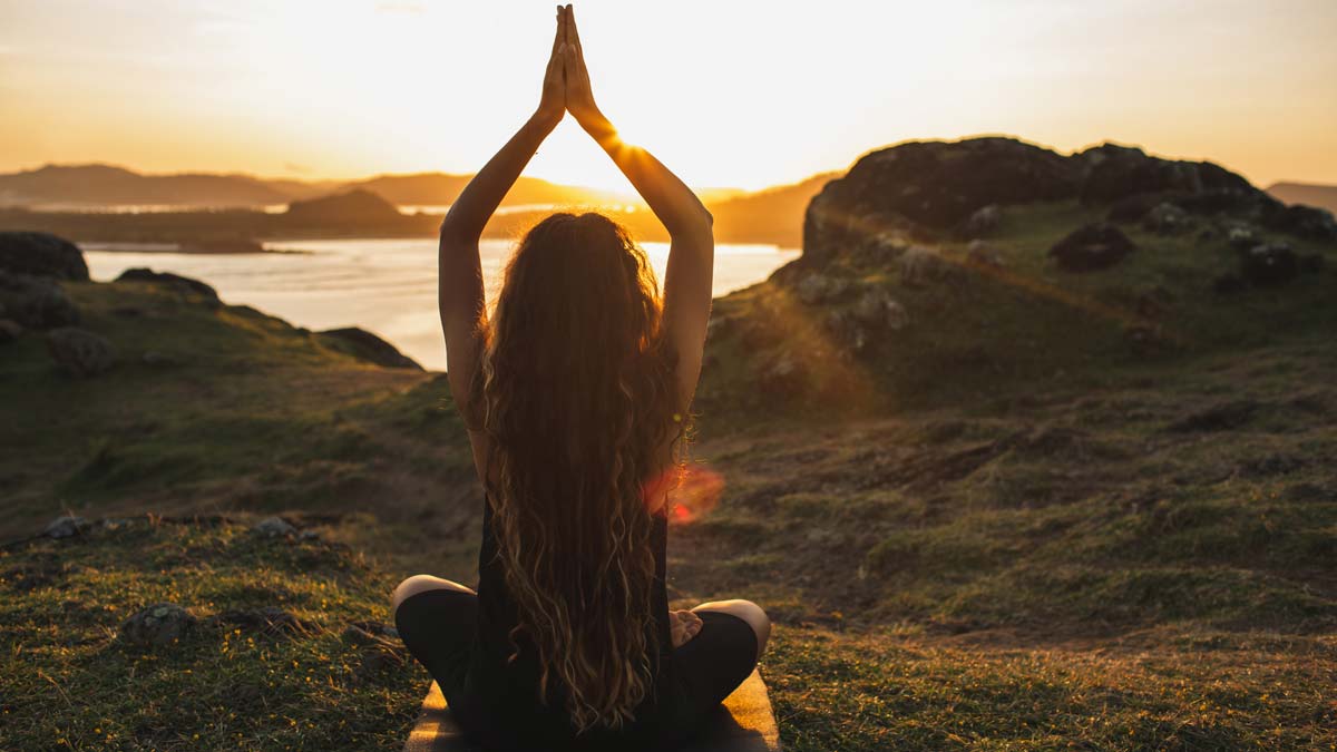 A woman with long wavy hair is sitting in a cross-legged yoga pose on a grassy hill overlooking water, hands raised above her head against a bright sunrise.