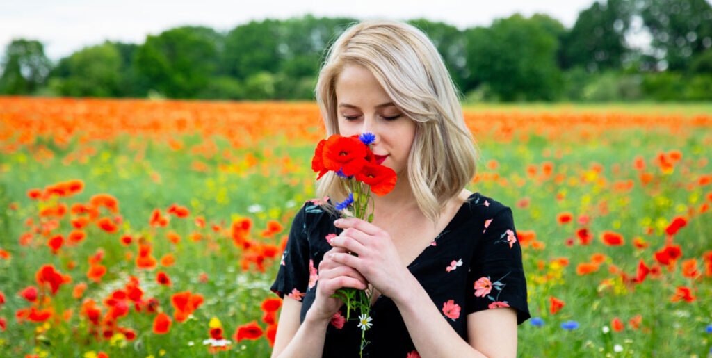 A blonde woman in a black floral dress in a vibrant red poppy field, smiling while smelling a small bouquet of poppies and cornflowers.