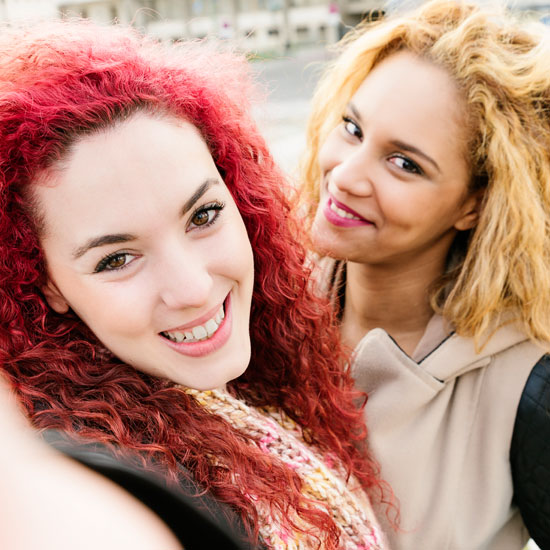 Two young women smiling and posing closely for a selfie; one has vibrant red curly hair, the other has curly blonde hair.