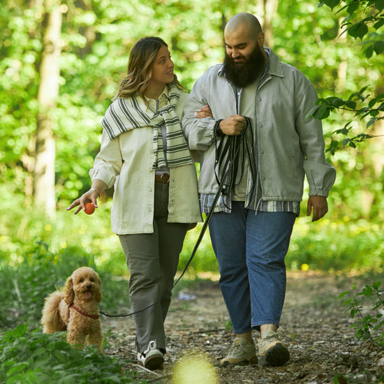 A man and a woman walk arm-in-arm on a wooded path with their small dog.