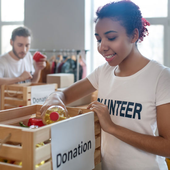 A smiling female volunteer packs a bottle of oil into a wooden crate labeled 'Donation' as part of a food drive.
