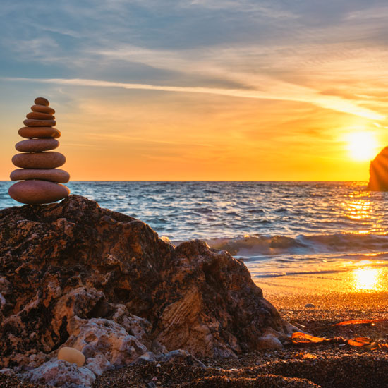 A perfectly balanced stack of smooth stones sits on a large rock on a beach at sunset, with golden light reflecting on the ocean and sand.