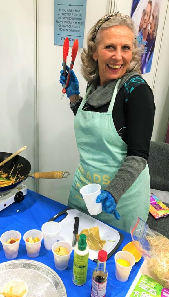 A member of staff poses with a healthy eating exhibition display. A small camping stove with a wok and various vegetables is on the table to her side.