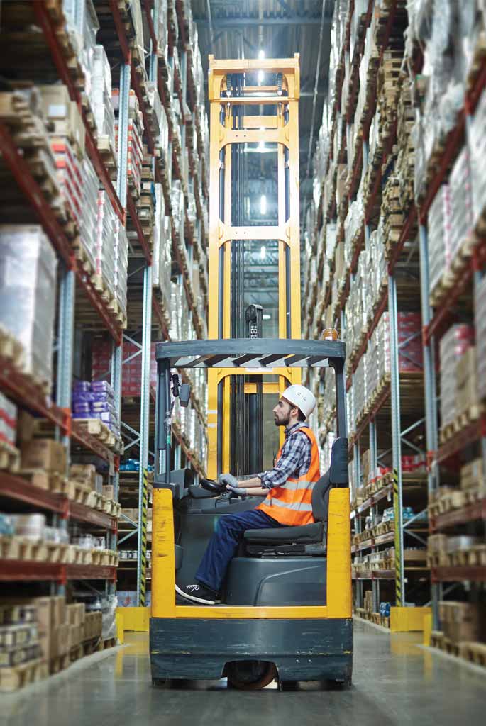 A man wearing a hard hat and safety vest operates a yellow forklift in a narrow warehouse aisle. The warehouse is huge, with very high shelves stretching far into the distance.
