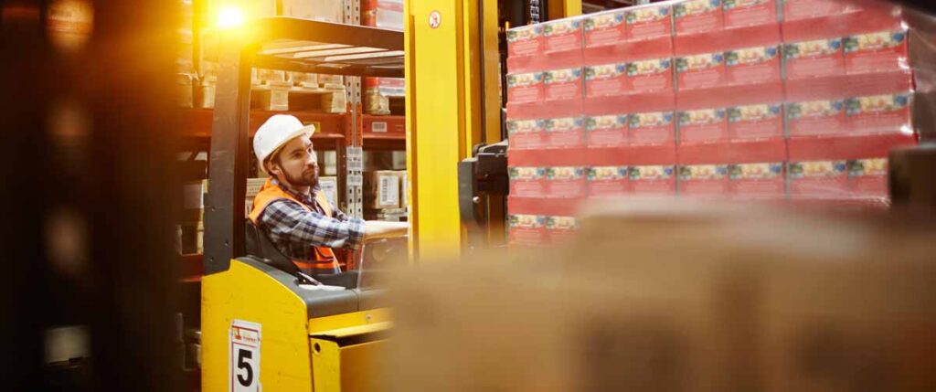 A warehouse worker in a white hard hat and orange safety vest is operating a yellow forklift carrying a large pallet of packaged goods. 
