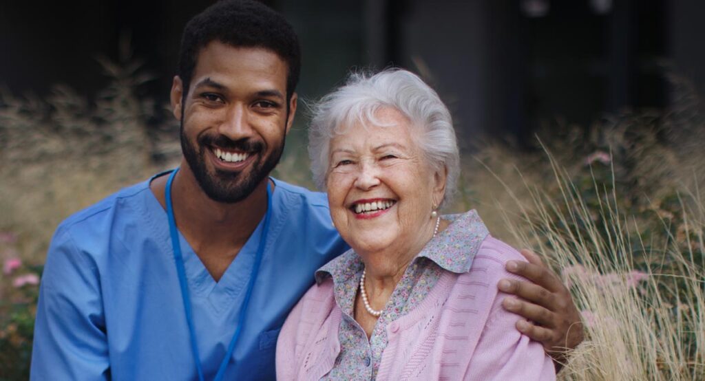 A young male caregiver in blue scrubs smiles happily with his arm around an elderly woman in a pink cardigan and pearl necklace; both are smiling.