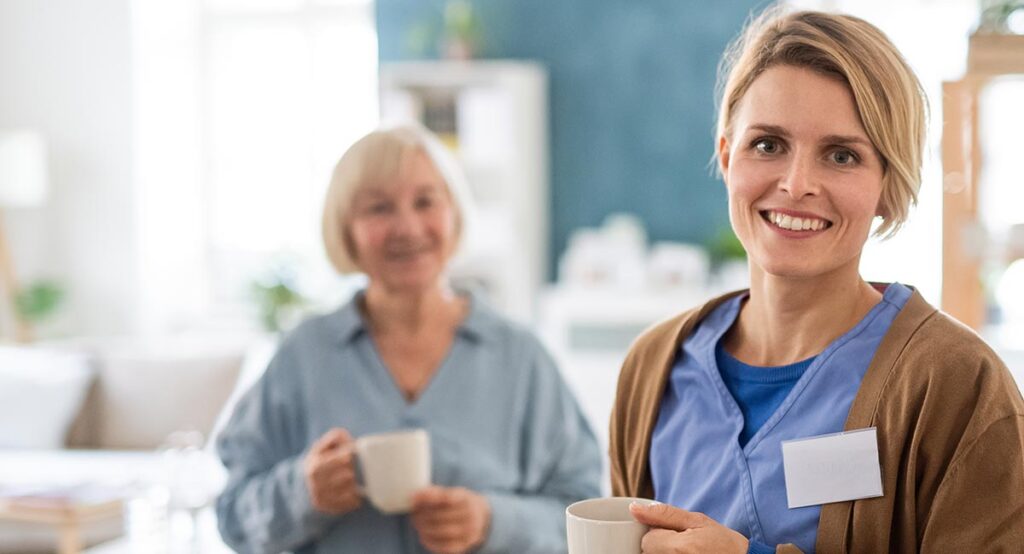 A young female caregiver smiles at the camera, holding a mug, with an older woman holding a mug in the background.