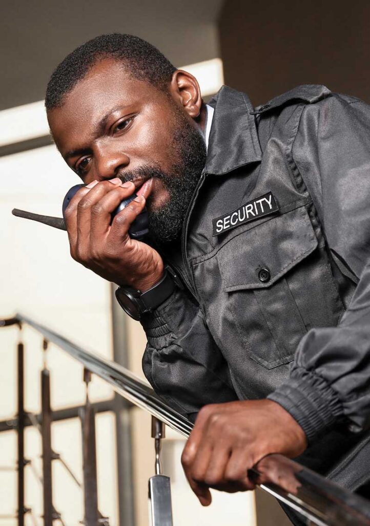 A male security guard in uniform leans on a railing indoors, speaking into a two-way radio.
