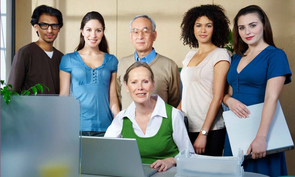 A diverse team of six colleagues, five standing and one seated at a desk with a laptop, smiling in an office environment.