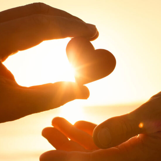 Two hands silhouetted against a golden sunset; one hand holds a small heart shape up to the light, passing it to the open hand below.