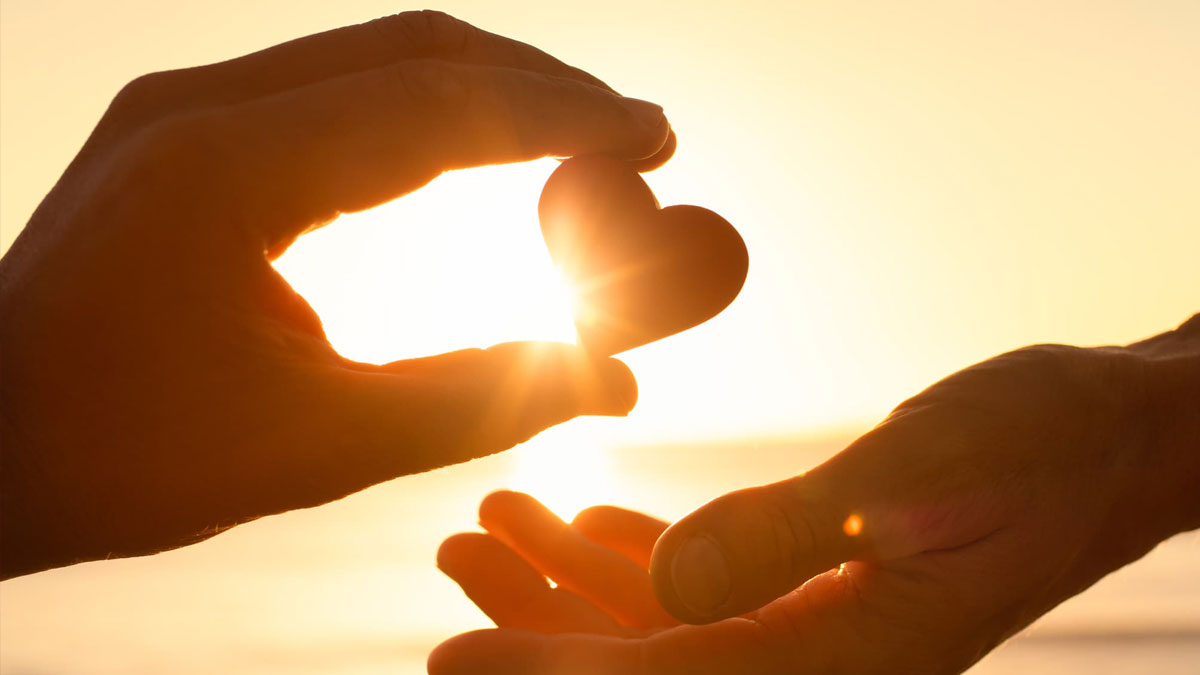 Two hands silhouetted against a golden sunset; one hand holds a small heart shape up to the light, passing it to the open hand below.