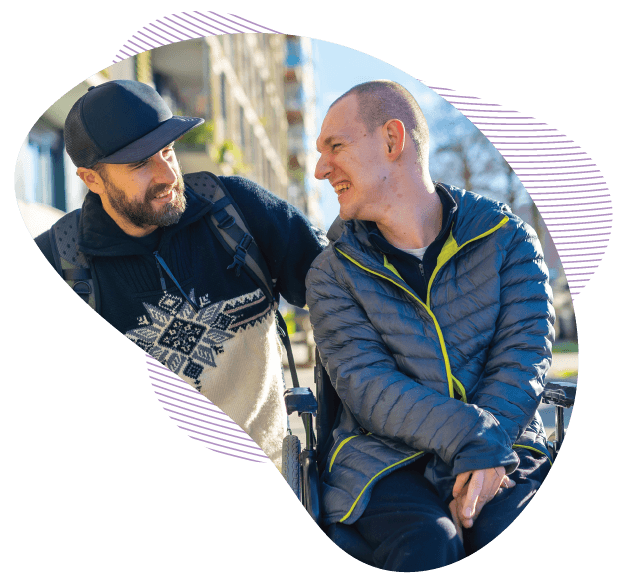 Two men share a laugh while talking outdoors on a sunny day; one man is seated in a wheelchair.