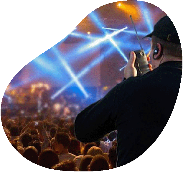 An event staff member in uniform uses a two-way radio while overseeing a vibrant night-time music festival crowd and brightly lit stage.