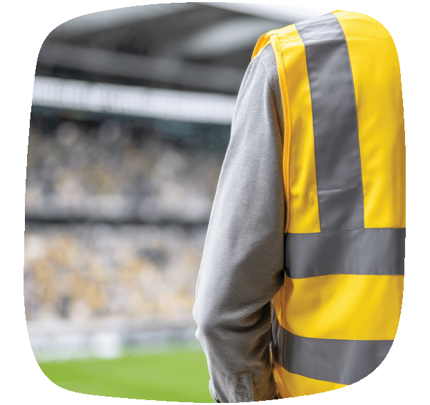 A person wearing a high-visibility yellow safety vest stands facing away from the camera in a stadium, with blurred green field and seating in the background.