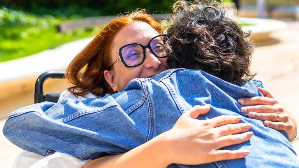 A smiling woman wearing glasses sits in a wheelchair and warmly embraces another person wearing a denim jacket outdoors.