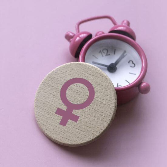 A small pink alarm clock sits next to a round wooden token featuring a pink female (Venus) symbol, set against a light pink background.