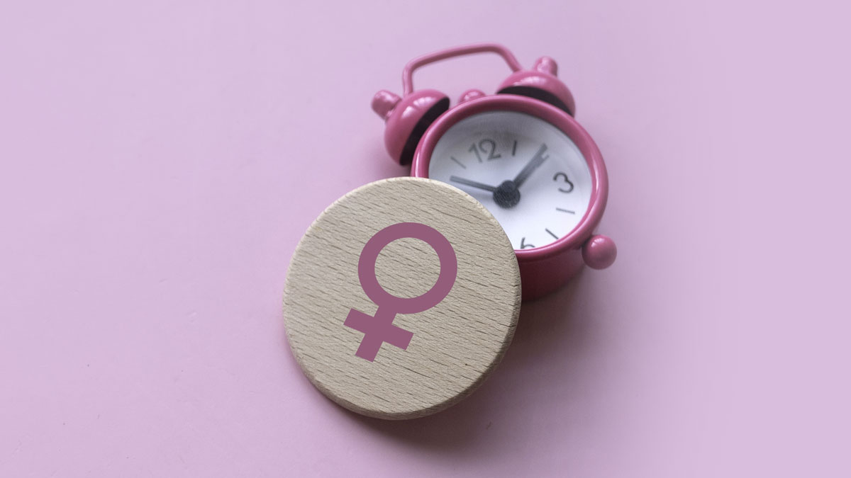 A small pink alarm clock sits next to a round wooden token featuring a pink female (Venus) symbol, set against a light pink background.