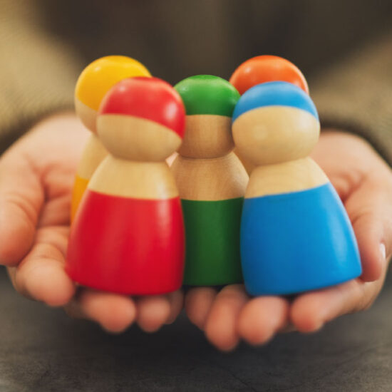 A person's hands gently cup and hold a small group of colourful wooden peg dolls.