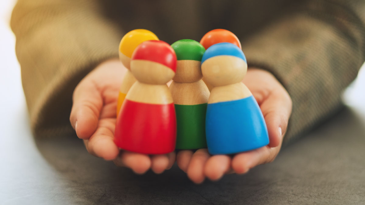 A person's hands gently cup and hold a small group of colourful wooden peg dolls.