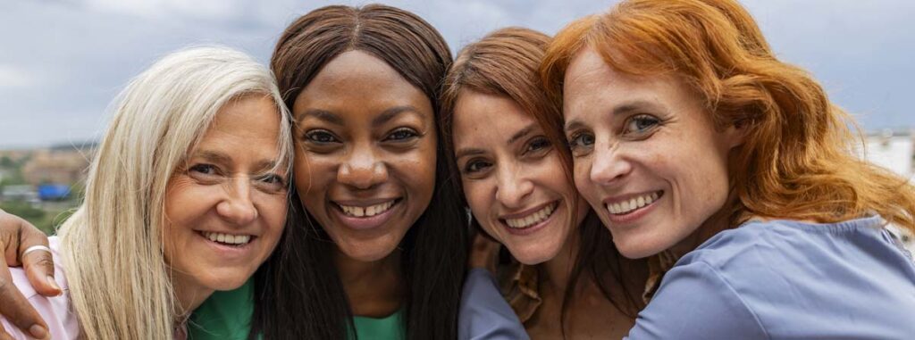 Four vibrant women of different ethnicities and ages huddled together smiling.