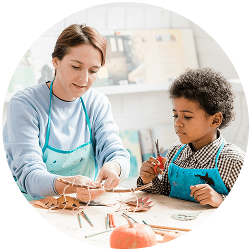 A woman and a young child wearing aprons are sitting at a table engaged in an arts and crafts activity, using natural materials and a small pumpkin.