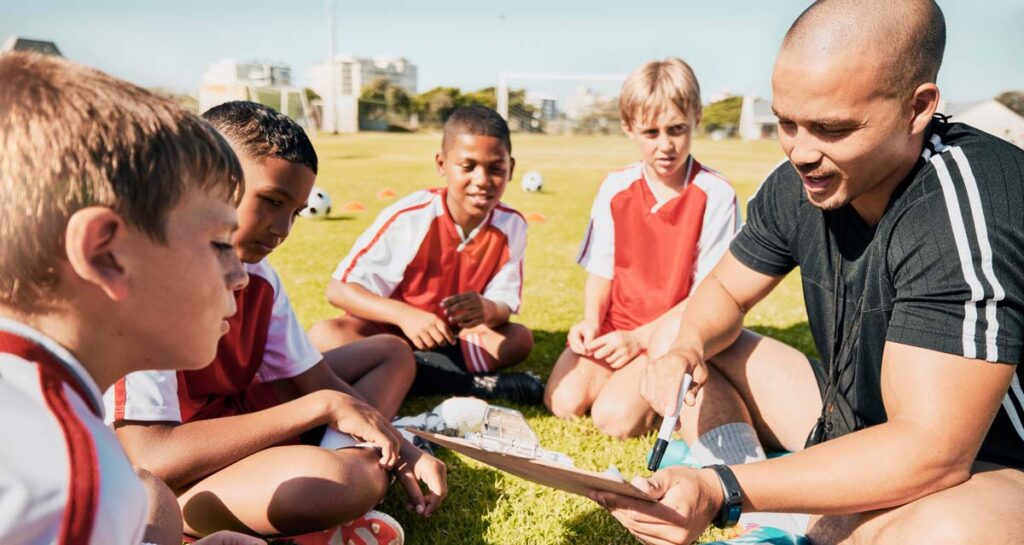 A male football coach sitting in a huddle with a team of young boys on a grassy field, reviewing strategy on a clipboard.