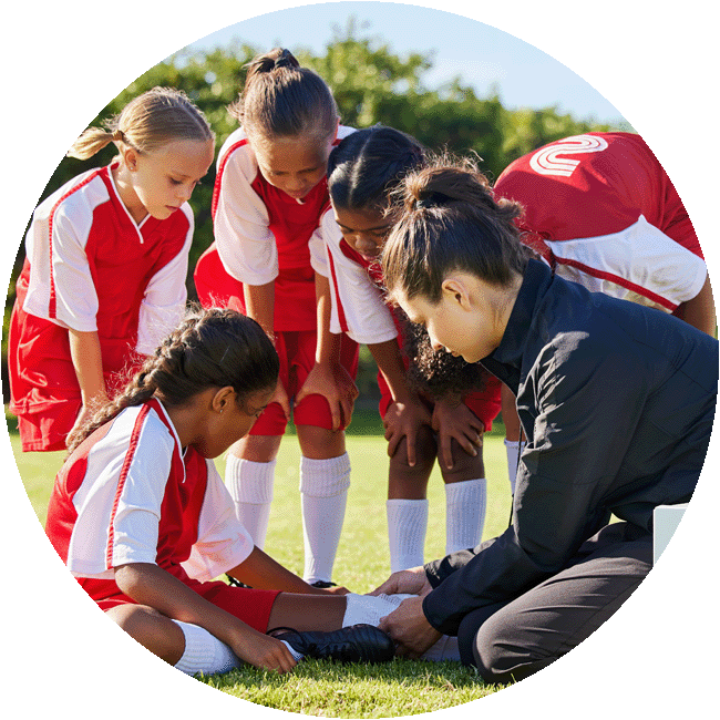 A female coach kneels on a football field, tending to the leg of a young player sitting down, while her worried teammates look on.