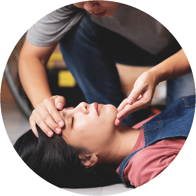 A man performs first aid by tilting back the head of an unconscious woman lying on the ground, checking for a pulse.
