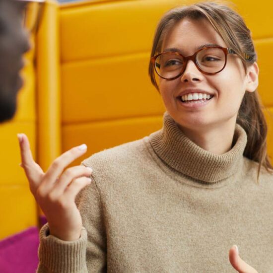 A young woman in large round glasses smiling, gesticulating with her hands, in conversation with a man who has his back to us.