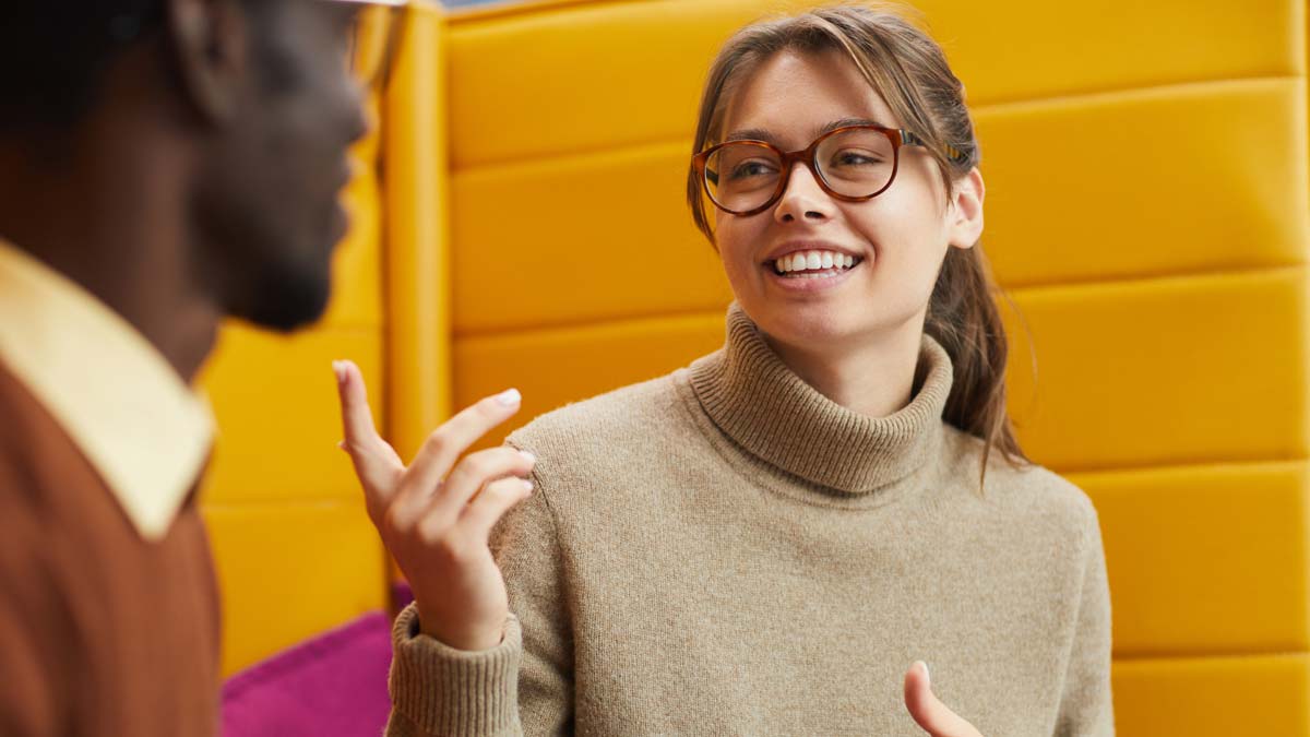A young woman in large round glasses smiling, gesticulating with her hands, in conversation with a man who has his back to us.