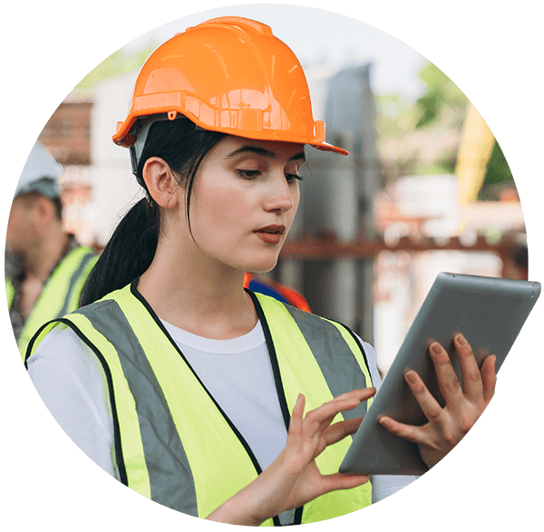 A female construction worker in an orange hard hat and high-visibility vest uses a digital tablet on a job site.