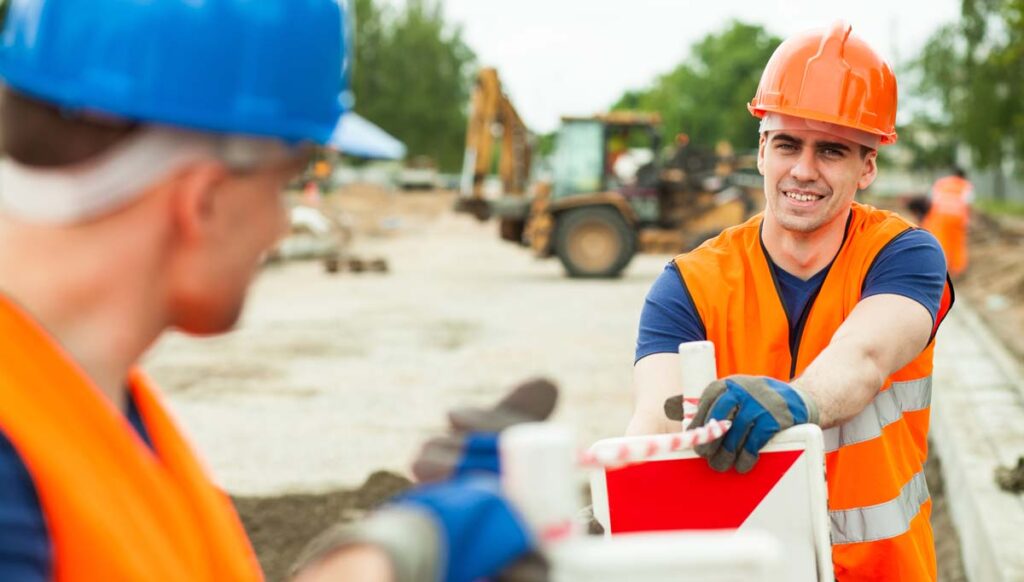 Two construction workers in safety gear are talking on a job site. Heavy machinery is visible in the background.