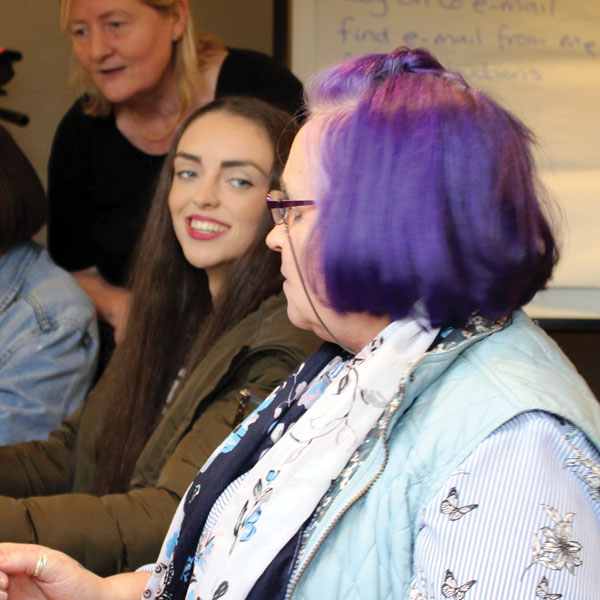 Two female learners using laptops: a young woman turns and grins broadly to an older woman with purple hair. A Crosby Training tutor stands behind them.
