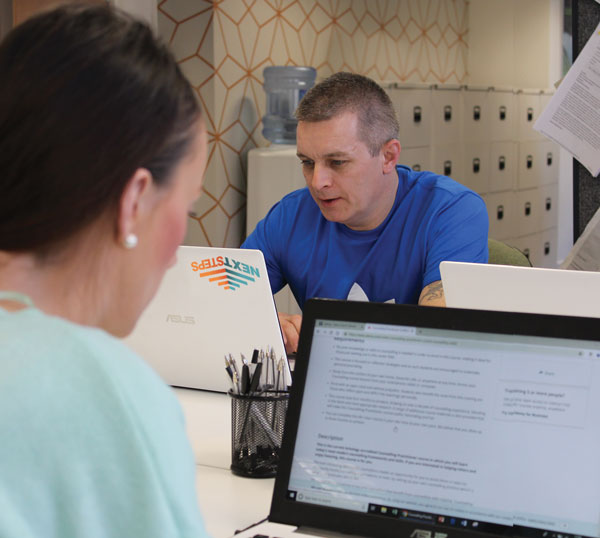 A male learner in our Next Steps Centre working on a laptop. In the foreground a female learner is viewing a website on a laptop.